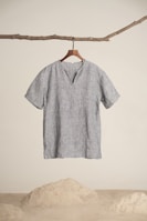 A simplistic short-sleeved, gray linen shirt hangs from a wooden hanger, attached to a natural branch. The background is minimalistic with a sandy floor and white wall.