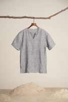 A simplistic short-sleeved, gray linen shirt hangs from a wooden hanger, attached to a natural branch. The background is minimalistic with a sandy floor and white wall.
