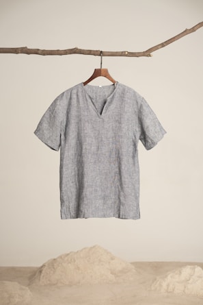 A simplistic short-sleeved, gray linen shirt hangs from a wooden hanger, attached to a natural branch. The background is minimalistic with a sandy floor and white wall.