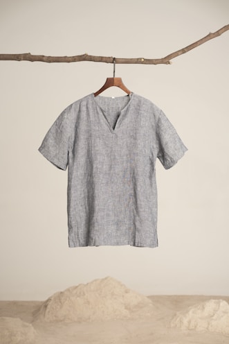 A simplistic short-sleeved, gray linen shirt hangs from a wooden hanger, attached to a natural branch. The background is minimalistic with a sandy floor and white wall.