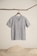 A simplistic short-sleeved, gray linen shirt hangs from a wooden hanger, attached to a natural branch. The background is minimalistic with a sandy floor and white wall.