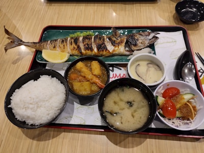 A traditional Japanese meal served on a tray, featuring a grilled whole fish with a lemon wedge, a bowl of white rice, a miso soup, a bowl with deep-fried items and vegetables in a sauce, a dish with chawanmushi (savory egg custard) topped with mushrooms, and a small salad with tomatoes and cucumbers.