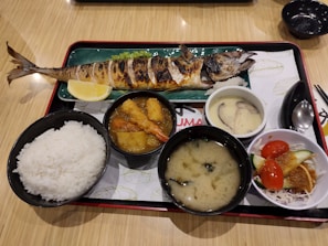 A traditional Japanese meal served on a tray, featuring a grilled whole fish with a lemon wedge, a bowl of white rice, a miso soup, a bowl with deep-fried items and vegetables in a sauce, a dish with chawanmushi (savory egg custard) topped with mushrooms, and a small salad with tomatoes and cucumbers.