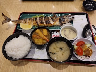 A traditional Japanese meal served on a tray, featuring a grilled whole fish with a lemon wedge, a bowl of white rice, a miso soup, a bowl with deep-fried items and vegetables in a sauce, a dish with chawanmushi (savory egg custard) topped with mushrooms, and a small salad with tomatoes and cucumbers.