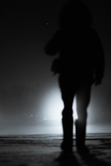 A silhouetted figure stands in the foreground against a backdrop of bright, diffused light. The setting appears to be dark and foggy, adding a mysterious and eerie atmosphere to the scene.