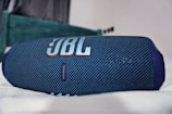 A blue portable Bluetooth speaker with a textured surface and the JBL logo prominently displayed in orange and white. It rests on a soft, white surface in a room with a blurred background.