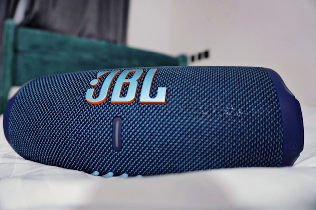 A blue portable Bluetooth speaker with a textured surface and the JBL logo prominently displayed in orange and white. It rests on a soft, white surface in a room with a blurred background.