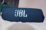 A portable Bluetooth speaker with a textured blue fabric exterior and a prominent logo displayed at the center. The speaker rests on a surface, with parts of a room in the softly blurred background.