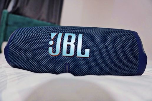 A portable Bluetooth speaker with a textured blue fabric exterior and a prominent logo displayed at the center. The speaker rests on a surface, with parts of a room in the softly blurred background.