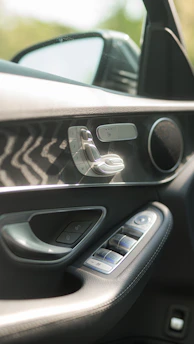 The interior panel of a car door featuring a metallic control panel with seat adjustment buttons and a speaker grille. The door handle is integrated into the sleek design, and the window controls are set into the leather trim. A side mirror is visible through the window, suggesting a focus on luxury and functionality.