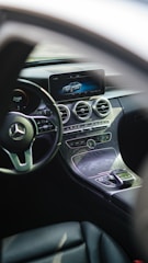 The interior of a luxury car featuring a sleek dashboard with a modern infotainment screen displaying a digital image of a car. The steering wheel prominently features a silver emblem. Below the screen, there are circular air vents and various controls embedded in a stylish, polished panel.