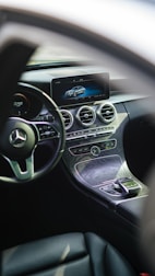 The interior of a luxury car featuring a sleek dashboard with a modern infotainment screen displaying a digital image of a car. The steering wheel prominently features a silver emblem. Below the screen, there are circular air vents and various controls embedded in a stylish, polished panel.
