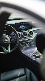 The interior of a luxury car featuring a sleek dashboard with a modern infotainment screen displaying a digital image of a car. The steering wheel prominently features a silver emblem. Below the screen, there are circular air vents and various controls embedded in a stylish, polished panel.