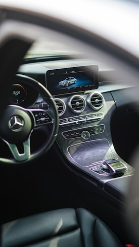 The interior of a luxury car featuring a sleek dashboard with a modern infotainment screen displaying a digital image of a car. The steering wheel prominently features a silver emblem. Below the screen, there are circular air vents and various controls embedded in a stylish, polished panel.