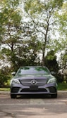 A luxury silver car is parked on a cobblestone path, surrounded by lush green trees. The car is centered in the frame, with its grille and emblem prominently displayed. The background features a mix of sunlight and shadows filtering through the tree branches.