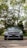 A luxury silver car is parked on a cobblestone path, surrounded by lush green trees. The car is centered in the frame, with its grille and emblem prominently displayed. The background features a mix of sunlight and shadows filtering through the tree branches.