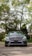 A luxury silver car is parked on a cobblestone path, surrounded by lush green trees. The car is centered in the frame, with its grille and emblem prominently displayed. The background features a mix of sunlight and shadows filtering through the tree branches.
