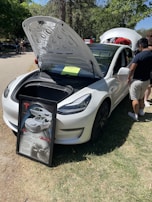 A white car with its front trunk open, revealing the storage compartment inside. In front of the car, a framed display board features images and text about the car. There are a few people standing near the car, with trees and greenery in the background.