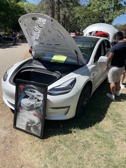 A white car with its front trunk open, revealing the storage compartment inside. In front of the car, a framed display board features images and text about the car. There are a few people standing near the car, with trees and greenery in the background.