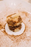 A dessert plated elegantly on a white dish, featuring a small, multi-layered cake with a crumbly texture. The cake appears to have layers of cream and a dusting of cocoa or cinnamon surrounds the plate, adding an artistic touch.