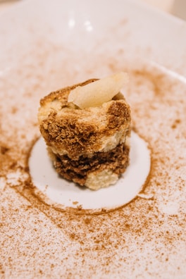 A dessert plated elegantly on a white dish, featuring a small, multi-layered cake with a crumbly texture. The cake appears to have layers of cream and a dusting of cocoa or cinnamon surrounds the plate, adding an artistic touch.