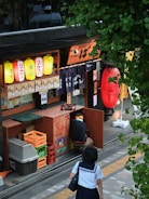 A street scene in front of a small Japanese restaurant with colorful paper lanterns hanging outside. A person in a school uniform walks by on a sidewalk, while another person kneels by storage boxes near the restaurant's entrance. Green foliage partially frames the scene from the top.