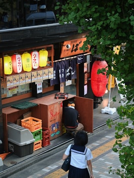 A street scene in front of a small Japanese restaurant with colorful paper lanterns hanging outside. A person in a school uniform walks by on a sidewalk, while another person kneels by storage boxes near the restaurant's entrance. Green foliage partially frames the scene from the top.