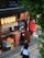 A street scene in front of a small Japanese restaurant with colorful paper lanterns hanging outside. A person in a school uniform walks by on a sidewalk, while another person kneels by storage boxes near the restaurant's entrance. Green foliage partially frames the scene from the top.