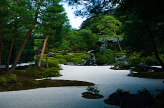 A serene Japanese garden features carefully manicured trees and bushes, a smooth gravel ground, and traditional stone elements. The garden is lush with green foliage and is partially enclosed by a modern glass building.