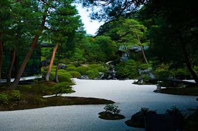 A serene Japanese garden features carefully manicured trees and bushes, a smooth gravel ground, and traditional stone elements. The garden is lush with green foliage and is partially enclosed by a modern glass building.