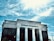 A bank building with white columns stands prominently under a bright blue sky with scattered clouds. The name 'First Citizens Bank' is clearly displayed on the facade of the building.