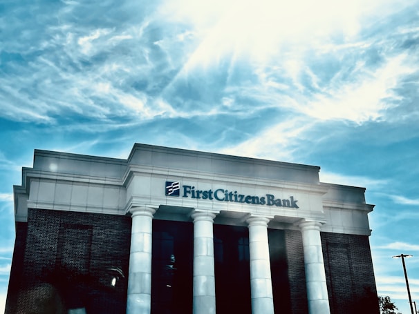A bank building with white columns stands prominently under a bright blue sky with scattered clouds. The name 'First Citizens Bank' is clearly displayed on the facade of the building.