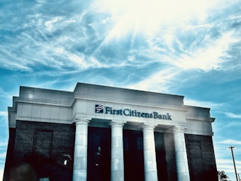 A bank building with white columns stands prominently under a bright blue sky with scattered clouds. The name 'First Citizens Bank' is clearly displayed on the facade of the building.