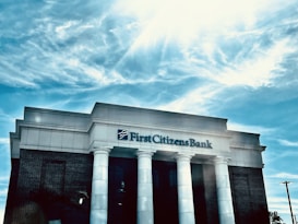 A bank building with white columns stands prominently under a bright blue sky with scattered clouds. The name 'First Citizens Bank' is clearly displayed on the facade of the building.