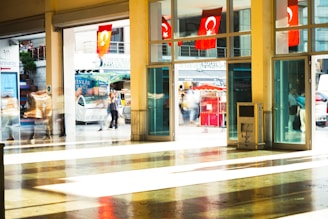 A busy indoor location with large windows showing multiple Turkish flags hanging outside. People are walking in both directions, their images blurred due to motion. The sunlight creates patterns on the shiny floor inside.