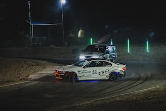 Two racing cars navigate through a dimly lit track at night. The foreground car, a sleek white vehicle with the number 45 and sponsor logos, is highlighted by blue and orange underglow lights. The background shows a rugged terrain vehicle, partially visible and slightly obscured by dust. Bright green accent lights line the course, illuminating the track's curves.
