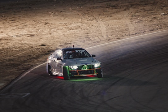 A racing car is captured in motion on a curving track at night. The car has various lights, including red and green underglow, and appears to be drifting as it maneuvers around the corner. The background is an arid, desert-like landscape with sandy terrain.