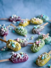 Assorted cake popsicles are arranged on a blue surface, each covered in vibrant colored chocolate coatings of purple, yellow, green, and adorned with white chocolate drizzle and mini marshmallows. Wooden sticks are used as handles for the popsicles.
