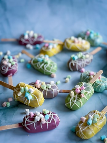 Assorted cake popsicles are arranged on a blue surface, each covered in vibrant colored chocolate coatings of purple, yellow, green, and adorned with white chocolate drizzle and mini marshmallows. Wooden sticks are used as handles for the popsicles.