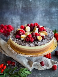A three-layered cake is elegantly presented with chocolate shavings on top, adorned with fresh raspberries, cherries, and toasted meringue pieces. The cake sits on a round, white platter, surrounded by scattered fruits and green leaves on a textured, dark surface.