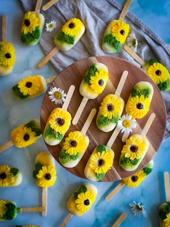 A collection of popsicles decorated with intricately crafted yellow flowers, resembling sunflowers, with green leaves. These popsicles are arranged both on a wooden circular board and scattered on a pastel blue and gray textured surface. Some small white daisy-like flowers are placed among the popsicles, enhancing the decorative appeal.