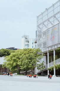 A group of children wearing red and black uniforms walk with an adult across a wide open area next to a modern building with a grid-like facade and large trees with green foliage. In the background, additional modern buildings are visible against a light blue sky.
