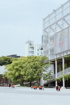 A group of children wearing red and black uniforms walk with an adult across a wide open area next to a modern building with a grid-like facade and large trees with green foliage. In the background, additional modern buildings are visible against a light blue sky.
