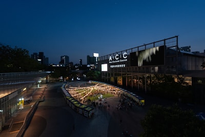 The scene captures an outdoor event at the Asia Culture Center during the evening. Strings of lights create a festive atmosphere over a gathering of people. Modern buildings illuminate the surroundings, while the sky transitions from daylight to twilight.
