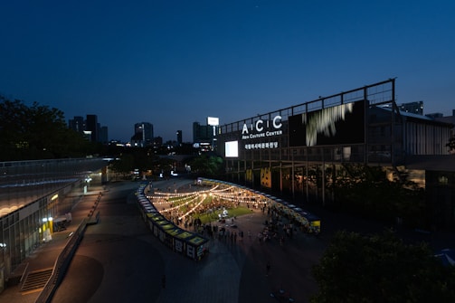 The scene captures an outdoor event at the Asia Culture Center during the evening. Strings of lights create a festive atmosphere over a gathering of people. Modern buildings illuminate the surroundings, while the sky transitions from daylight to twilight.