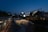 The scene captures an outdoor event at the Asia Culture Center during the evening. Strings of lights create a festive atmosphere over a gathering of people. Modern buildings illuminate the surroundings, while the sky transitions from daylight to twilight.