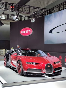 A sleek, red Bugatti sports car is showcased under bright lights on a platform. The car features glossy black accents and aerodynamic design elements. A large Bugatti logo is displayed on a nearby wall, and a person stands near the car in the background.