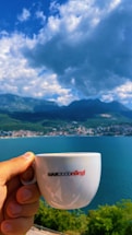 A hand holds a white coffee cup labeled 'BAR2000ellegi' against a scenic background. The setting includes a large body of water, a distant town along the shoreline, and a range of mountains under a partly cloudy sky.