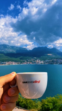 A hand holds a white coffee cup labeled 'BAR2000ellegi' against a scenic background. The setting includes a large body of water, a distant town along the shoreline, and a range of mountains under a partly cloudy sky.