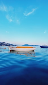 A small white boat floating in calm blue water, with gentle ripples reflecting the sky. A scenic view of a coastal landscape is visible in the background along with a larger boat on the right side. The sky is clear with some wispy clouds.